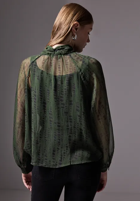 Frau mit grün gemusterter, transparenter Bluse von hinten, Licht von links.